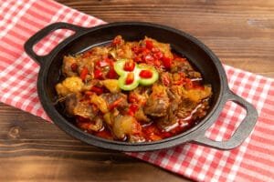 Sajian karahi daging atau ayam dengan tomat dan cabai merah dalam wajan bulat hitam, diletakkan di atas taplak meja kotak-kotak.