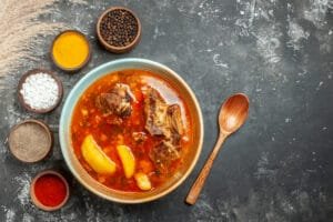 Semangkuk sup daging kental berwarna merah, kemungkinan Nihari, dengan potongan daging dan kentang, ditemani sendok kayu dan mangkuk berisi rempah-rempah di sekelilingnya.